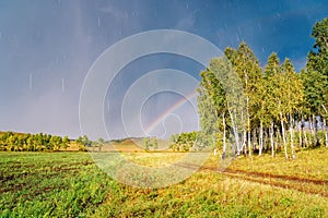 The autumn forest and fields are lit by the sun after the rain, and the sky is covered with clouds and a rainbow
