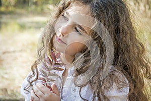 In the autumn forest, a small beautiful girl with her hair is holding a rowan leaf in her hands