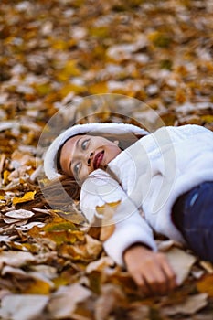 Autumn girl playing in city park. Portrait of an autumn woman lying over leaves and smiling outside in fall forest. Beautiful ener