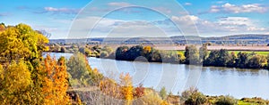 Autumn landscape with a forest on a hill near the river and a picturesque blue sky with white clouds in sunny weather