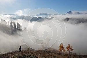 Autumn landscape of high mountains, orange coloured trees, fog. Sun rays enlighten the lawn with dry grass. Blue sky.