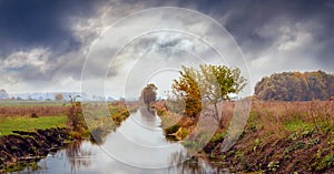 Autumn landscape with a river in the field, trees on the shores and a dramatic stormy sky. Autumn view in cloudy weather