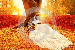 Autumn Landscape. Romantic Victorian Style Woman reading Book in Park with Orange Maple Leaves. Colorful Fall Season Bright