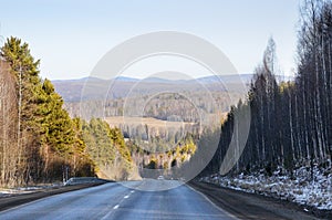 Autumn landscape with tarmac road on the smooth hills covered with taiga forest under blue sky