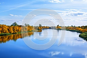 Autumn landscape, yellow leaves trees on river bank on blue sky and white clouds background on sunny day, reflection in blue water