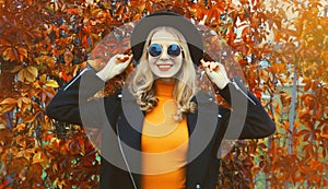 Autumn portrait of beautiful happy smiling young woman wearing black round hat, sunglasses and coat jacket in the park on leaves