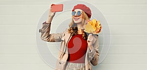 Autumn portrait of beautiful smiling woman taking selfie picture by phone with yellow maple leaves wearing red french beret