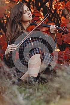 Autumn portrait of beautiful woman sitting on the ground with a violin under chin on a background of red foliage, girl engaged in