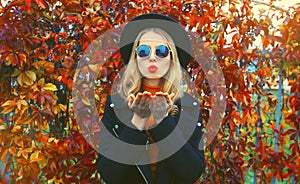 Autumn portrait of beautiful young woman blowing her lips sends sweet air kiss wearing black round hat, sunglasses and coat jacket