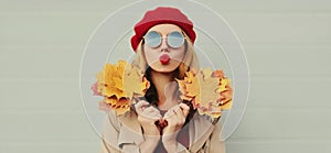 Autumn portrait beautiful young woman model with yellow maple leaves blowing her lips with lipstick wearing a red french beret