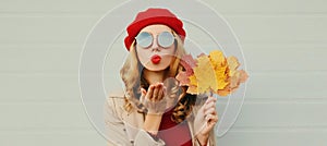 Autumn portrait beautiful young woman model with yellow maple leaves blowing her lips with lipstick wearing a red french beret