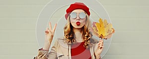 Autumn portrait beautiful young woman model with yellow maple leaves blowing her lips with lipstick wearing a red french beret