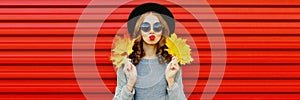 Autumn portrait of beautiful young woman posing with yellow maple leaves blowing her lips wearing black round hat, gray knitted