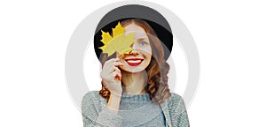 Autumn portrait happy smiling young woman covering her eye with yellow maple leaves wearing a black round hat, gray knitted