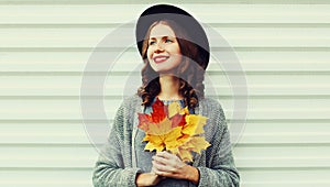 Autumn portrait happy smiling young woman with yellow maple leaves looking away wearing a black round hat, gray knitted sweater