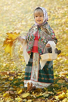 Autumn portrait of the little girl in the traditional russian sarafan and headscarf gathering yellow leaves and pinecones