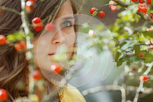 Autumn portrait of pensive young woman behind the branches of a rosehip bush with red berries, beautiful girl face with a pensive