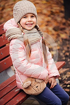 Autumn portrait of smiling child girl sitting on bench in park in warm knitted hat