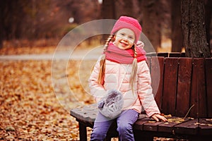 autumn portrait of smiling child girl sitting on bench in park in warm knitted hat