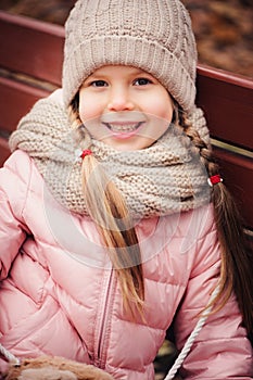 Autumn portrait of smiling child girl sitting on bench in park in warm knitted hat and scarf