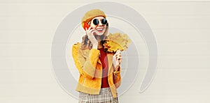Autumn portrait smiling woman calling on phone holding yellow maple leaves wearing french beret on city street over gray wall