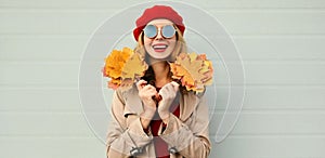 Autumn portrait of smiling young woman with yellow maple leaves wearing red french beret over gray