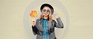 Autumn portrait of stylish happy smiling young woman with yellow maple leaves wearing round hat and rock style black leather