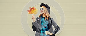 Autumn portrait of stylish happy smiling young woman with yellow maple leaves wearing round hat and rock style black leather