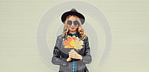 Autumn portrait of stylish happy smiling young woman with yellow maple leaves wearing round hat and rock style black leather