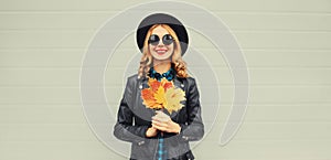 Autumn portrait of stylish happy smiling young woman with yellow maple leaves wearing round hat and rock style black leather