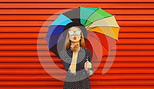 Autumn portrait of stylish young woman posing with colorful umbrella wearing black round hat blowing her lips sends kiss on red