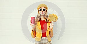 Autumn portrait woman with yellow maple leaves, coffee cup blowing red lips sending sweet air kiss over gray wall