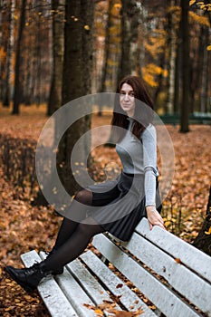 Autumn portrait of young woman in autumn park, sitting on the back of a white bench and enjoying the fall landscape