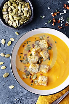 Autumn pumpkin vegetable cream soup in bowl served with seeds and crouton on black stone table from above