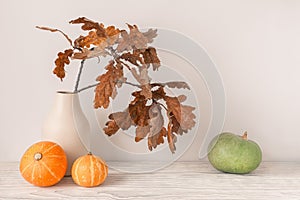 Autumn season concept. Dry oak leaves in vase with decorative pumpkins on light wooden table