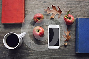 Autumn season still life with red apples, books, mobile device, black coffee cup and fall leaves over rustic wooden background. Kn