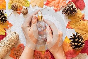 Autumn spa concept with essential oil bottle in female hands, dried leaves, pine cones on grey background. Organic cosmetic