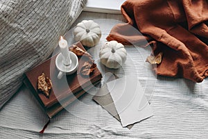 Autumn still life. Burning candle, books and linen pillow near window. Blank greeting card mockup scene. Moody