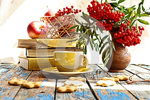 Autumn still life: a cup of tea with cookies scattered on a wooden table, against the backdrop of a stack of books and an autumn