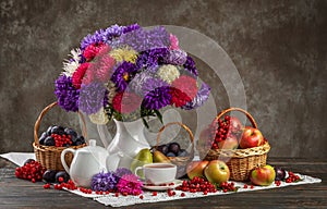 Autumn still life. A vase with asters, baskets of fruit, apples and plums and porcelain teaware on the table