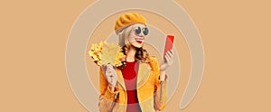 Autumn style outfit, portrait of fashionable smiling young woman model with mobile phone holds yellow maple leaves wearing orange