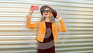 Autumn style outfit, stylish elegant happy woman posing taking selfie with mobile phone wearing hat, orange jacket and sunglasses