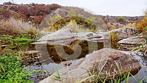 Autumn trees and large stone boulders around. A water cascade in autumn creek with fallen leaves. Water flows around the