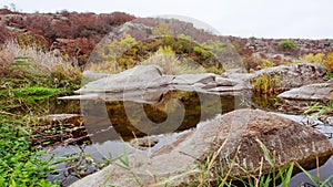 Autumn trees and large stone boulders around. A water cascade in autumn creek with fallen leaves. Water flows around the