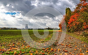 Autumn windy day in Germany, cloudy sky yellow leaves falling from trees standing near a dirt road and farmland. In the distance,