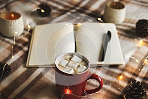 Autumn and winter background with notebook, pen, garland, cones, mug of cocoa, coffee or hot chocolate with marshmallow on warm pl