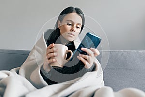 Autumn winter portrait of a young girl resting at home on the sofa with mobile phone and cup of hot drink, under warm blanket