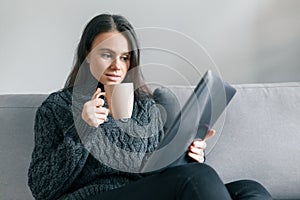 Autumn winter portrait of young girl in warm knitted sweater at home on the sofa with a digital tablet and cup of hot drink