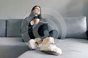 Autumn winter portrait of young girl in warm knitted sweater at home on the sofa with a digital tablet and cup of hot drink