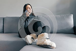 Autumn winter portrait of young girl in warm knitted sweater, at home on the sofa with a digital tablet and phone